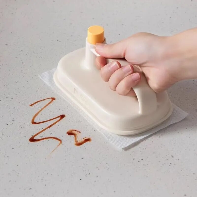 Hand using a white cleaning tool with an orange handle to wipe up a brown stain on a light surface.